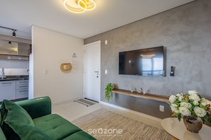 Een moderne woonkamer met een groene bank, een flatscreen televisie bevestigd aan een gestructureerde muur, een klein houten plankje met decoratieve items, en een bloevas.