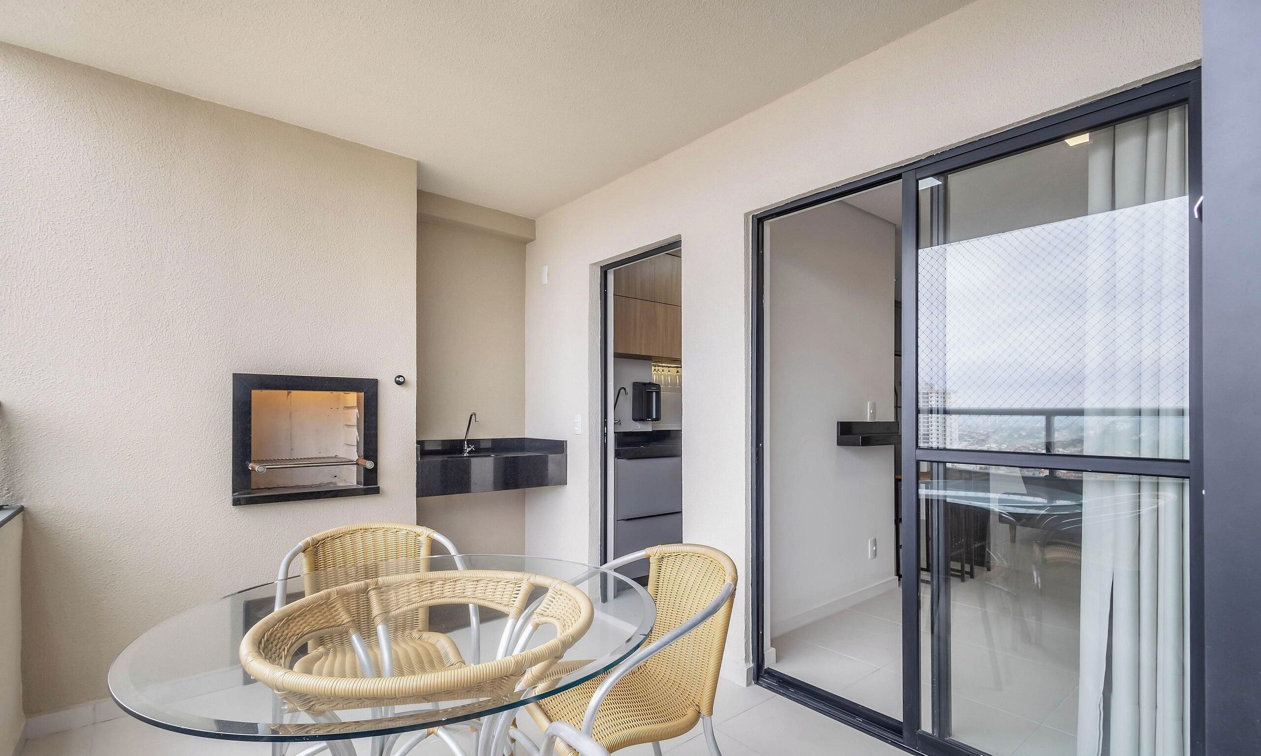 Un balcón con sillas de mimbre y una mesa de vidrio, una kitchenette con microondas y un amplio ventanal corredera que da a un espacio exterior.