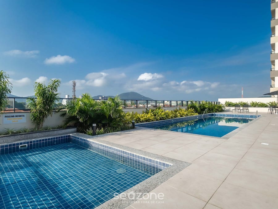 A rooftop swimming pool with tiled edges, surrounded by greenery and a clear sky.