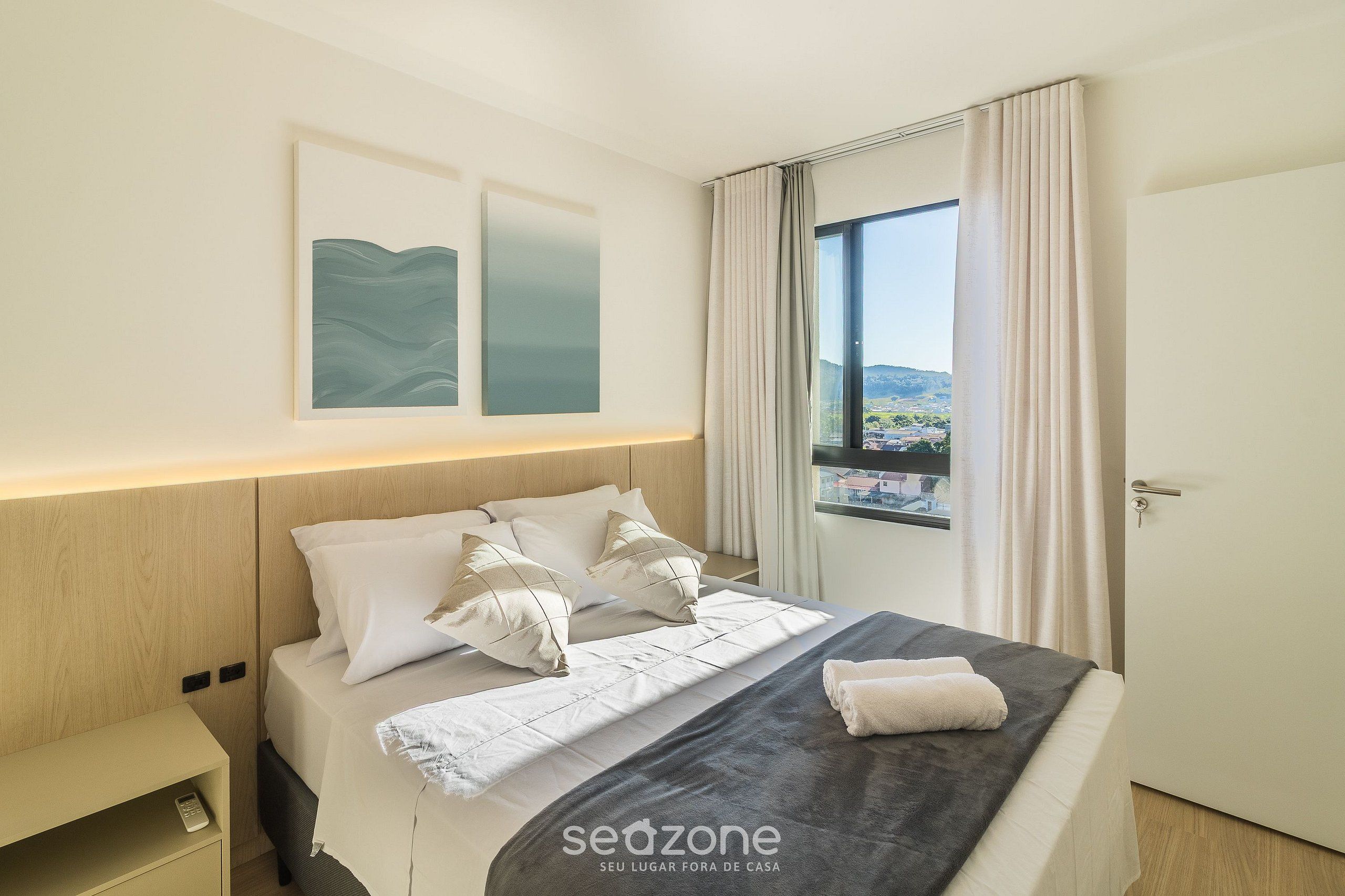 Una habitación de hotel con cama, mesita de noche, ventana con cortinas y una obra de arte en la pared.