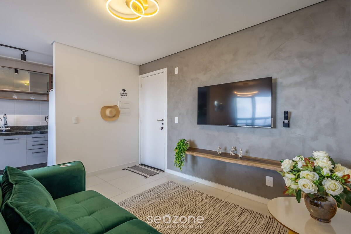 Een moderne woonkamer met een groene bank, een flatscreen televisie bevestigd aan een gestructureerde muur, een klein houten plankje met decoratieve items, en een bloevas.