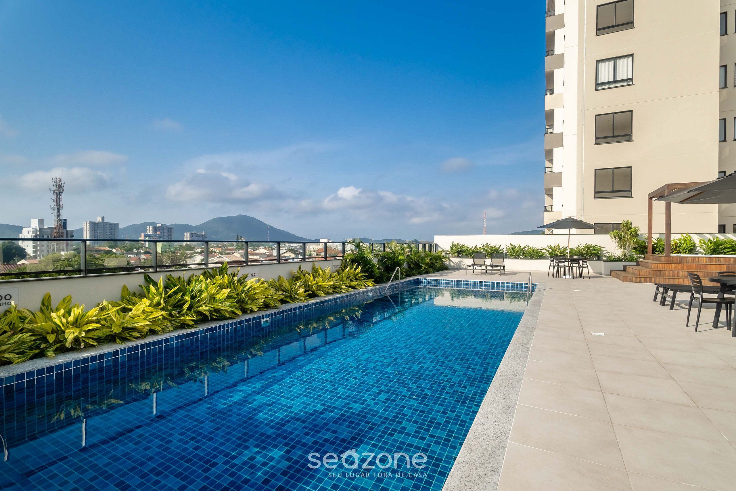 A rooftop pool with tiled flooring, surrounded by greenery and outdoor furniture.