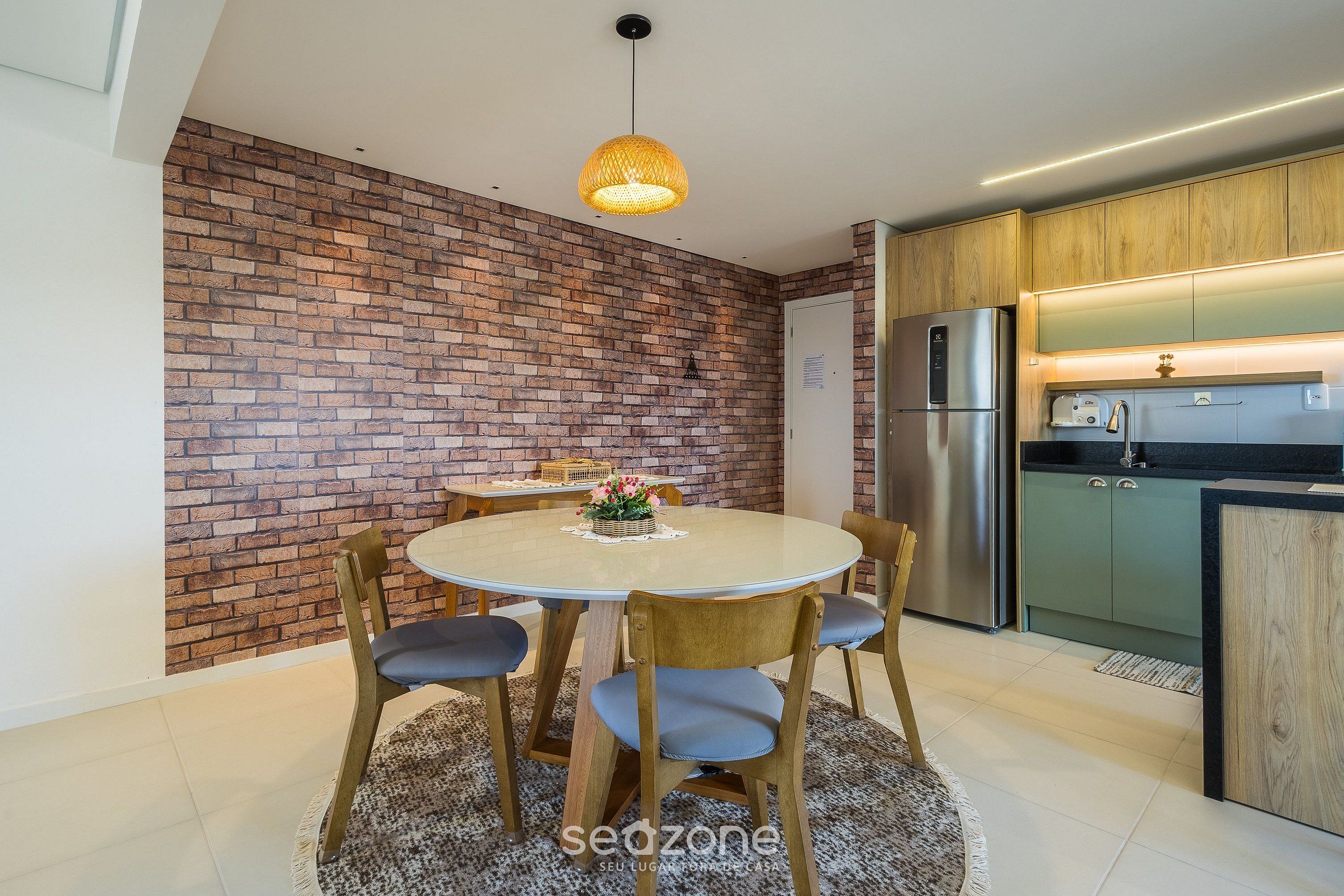 A modern kitchen with a brick wall, a dining area with a round table and chairs, and a refrigerator.