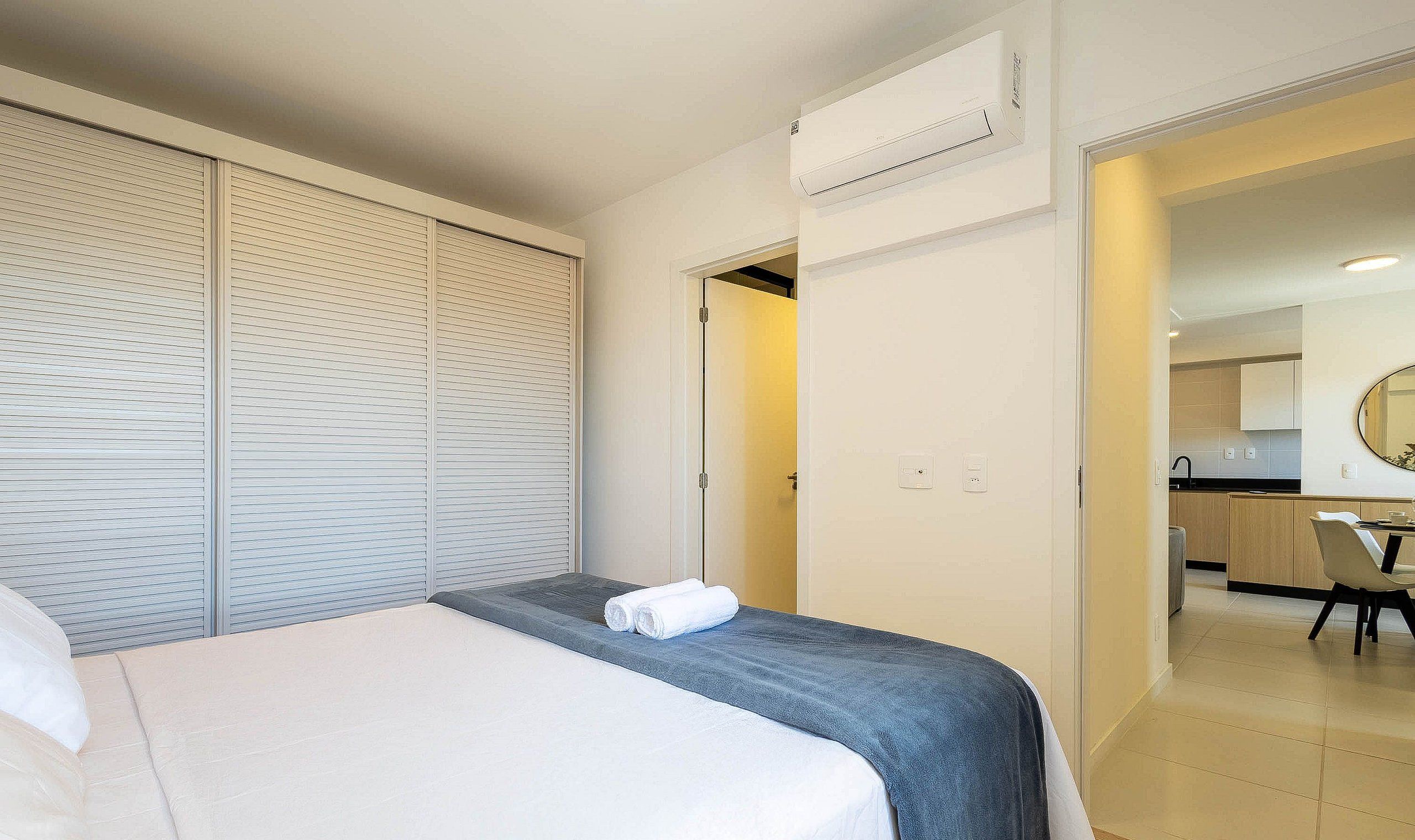 A hotel room with a bed, a wardrobe, an air conditioning unit, and a view into the kitchen area.