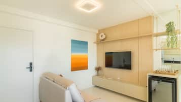 A modern living room with a flat-screen TV, a sofa, a painting, and a small kitchen area with a refrigerator.
