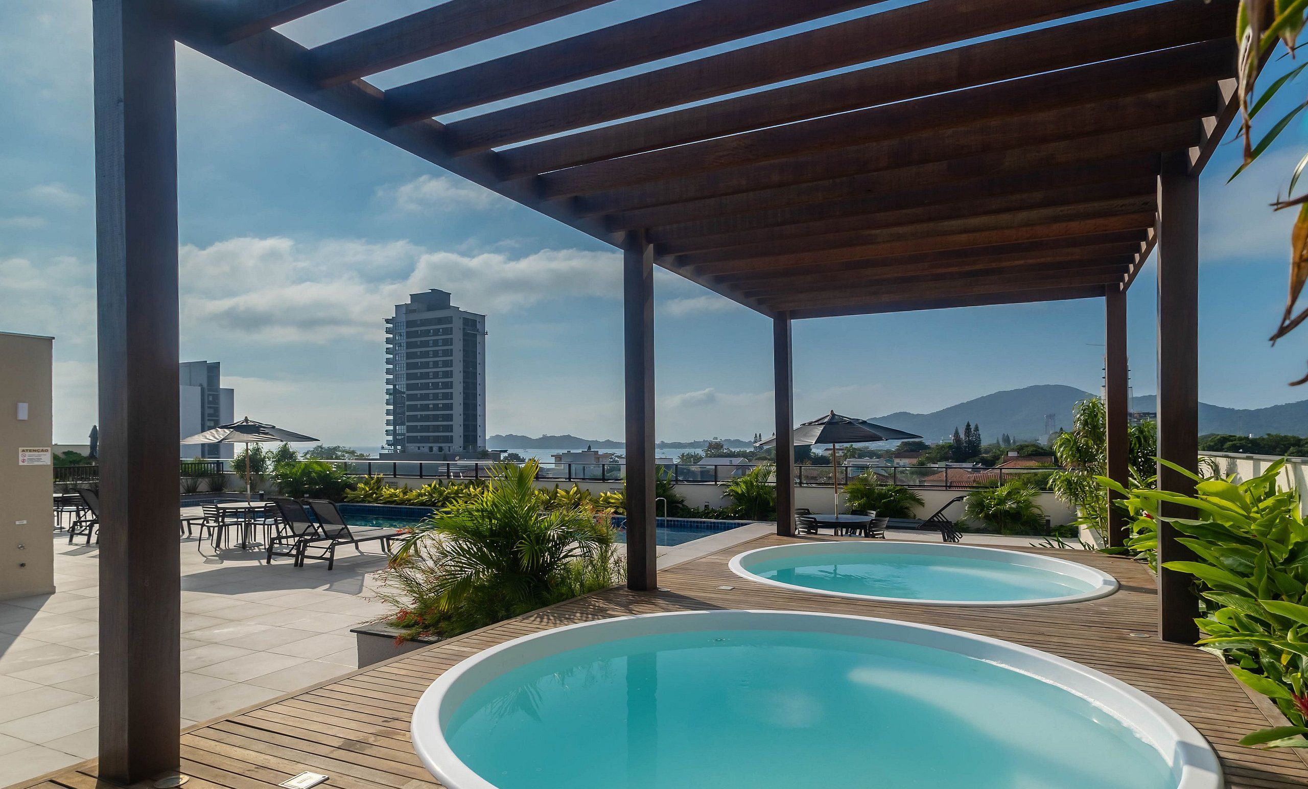 Un área de piscina en la azotea con dos jacuzzis, pérgolas de madera y vistas a la ciudad y montañas.
