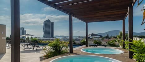 A rooftop pool area with two hot tubs, wooden pergolas, and a view of a cityscape and mountains.