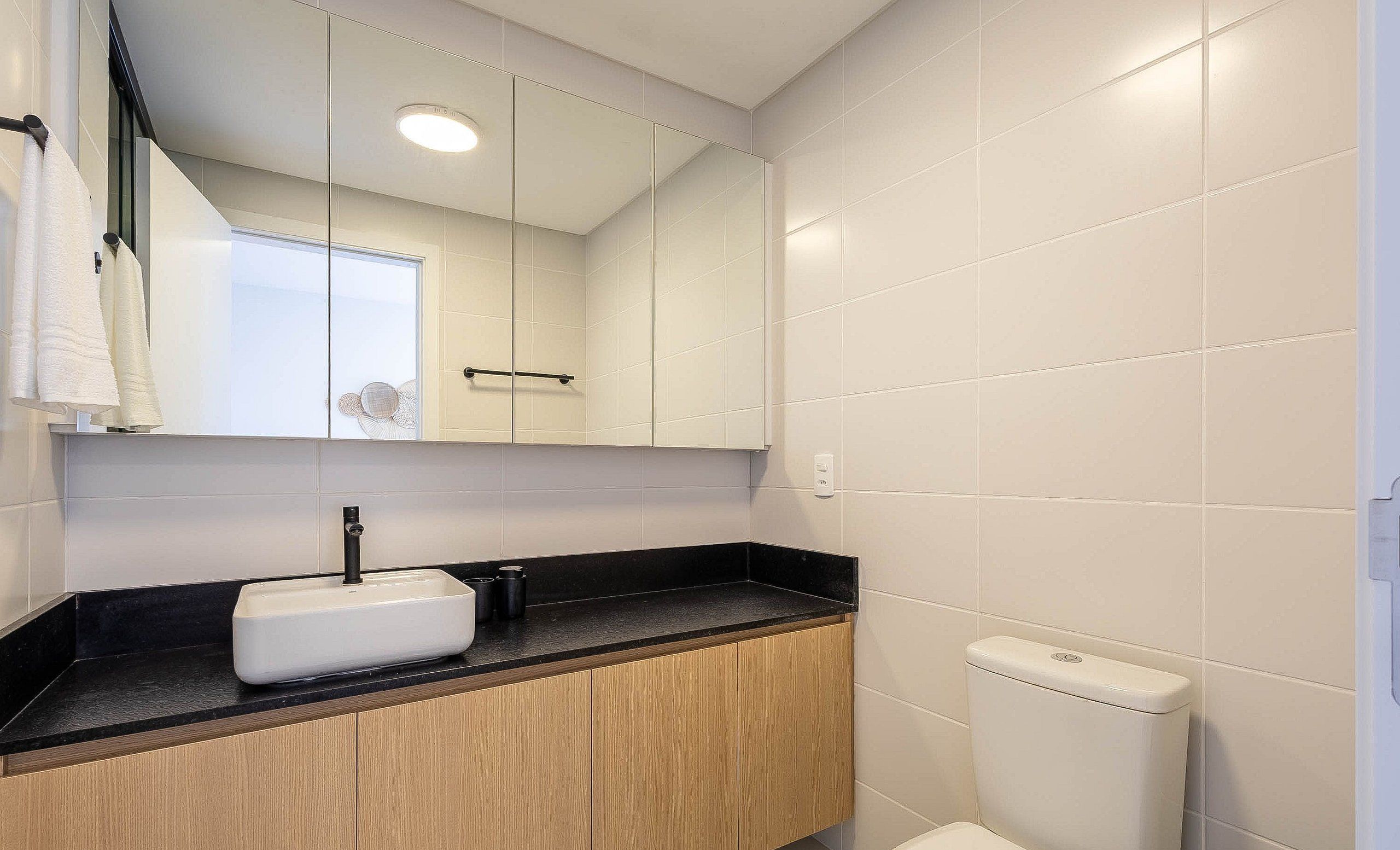 A bathroom with white tiles, a black countertop, a wooden cabinet, a white sink, a mirror, and a toilet.