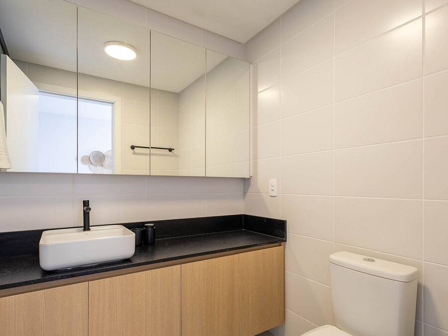 Ein Badezimmer mit weißen Fliesen, einer schwarzen Arbeitsplatte, einem Holzkabinet und einem weißen Waschbecken, einem Spiegel und einer Toilette.