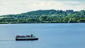 A boat with passengers on a calm river, surrounded by lush greenery and a distant building.