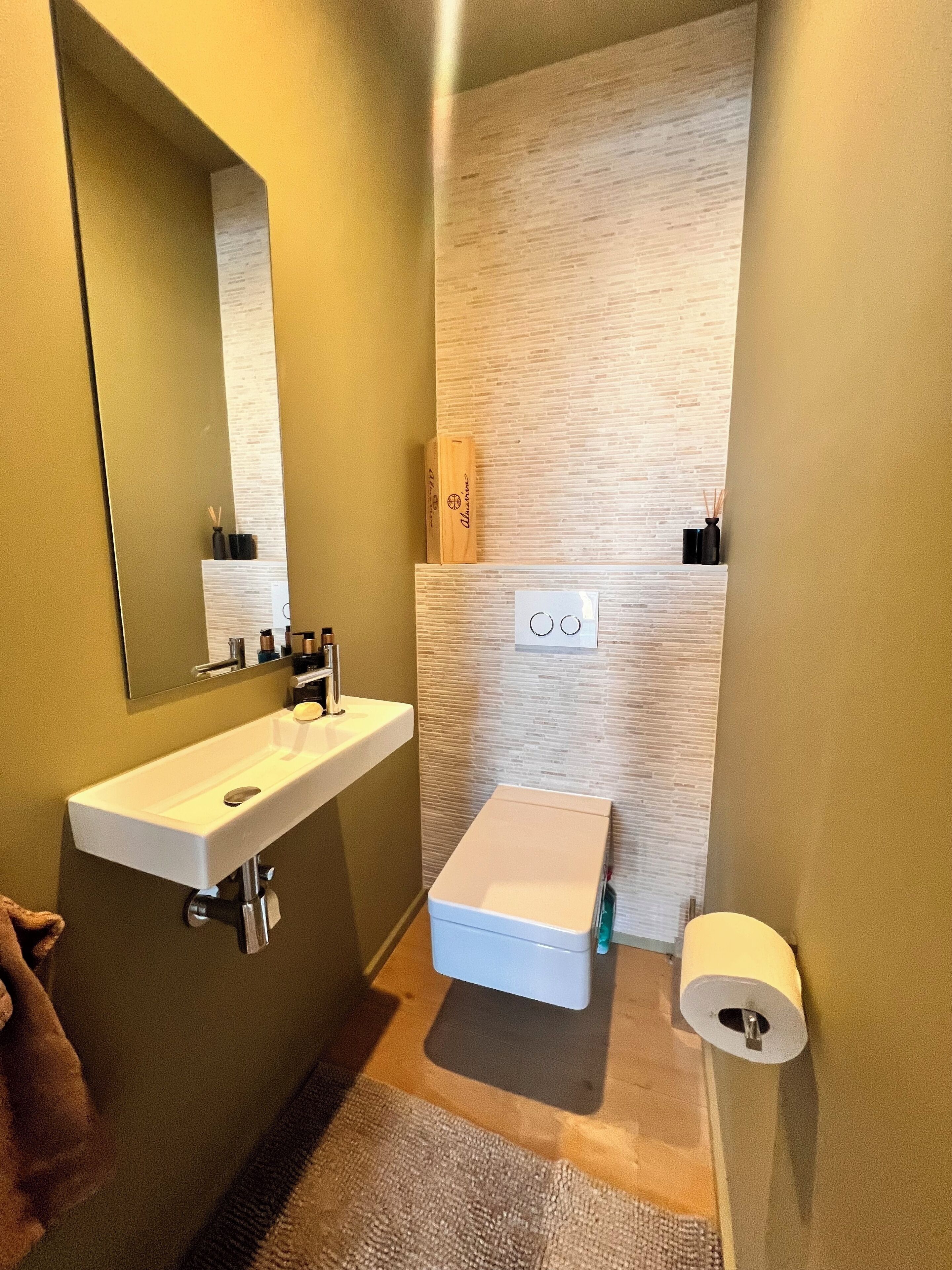 A bathroom with a white sink, mirror, and toilet. There is a towel on the floor and a roll of toilet paper on the wall.