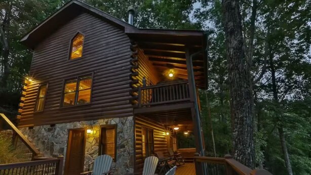 A two-story log cabin with a stone foundation, a balcony with seating, and a wooden deck.
