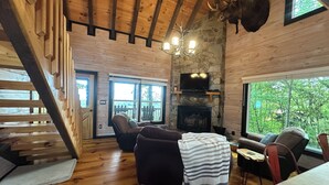 A cozy cabin interior with a stone fireplace, wooden floors, a mounted moose head, and a large window offering a view of the outdoors.