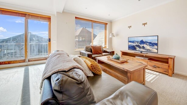A modern living room with a sofa, coffee table, and TV, large windows with blinds, and a mountain view. - Schnapps 3 - Mt Hotham (Hotham Heights)