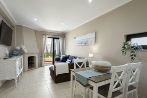A living room with a fireplace, a sofa, a dining table with chairs, a TV mounted on the wall, and a balcony with a view. - Hopstays - Forte Beach Retreat (Albufeira)