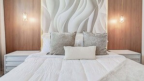 Un dormitorio de diseño moderno, con una cama grande vestida con ropa de cama blanca y varios almohadones, un panel decorativo en la pared y dos lámparas de noche.