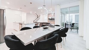 Una cocina moderna con una isla central, un comedor con una mesa larga y sillas, y una escalera que lleva a un nivel superior.