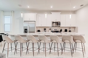A modern kitchen with white cabinets, a central island with bar stools, a refrigerator, microwave, and a coffee machine. - 9140 Mario Game Room, Theater & Pool! Solara 6BR (Kissimmee)