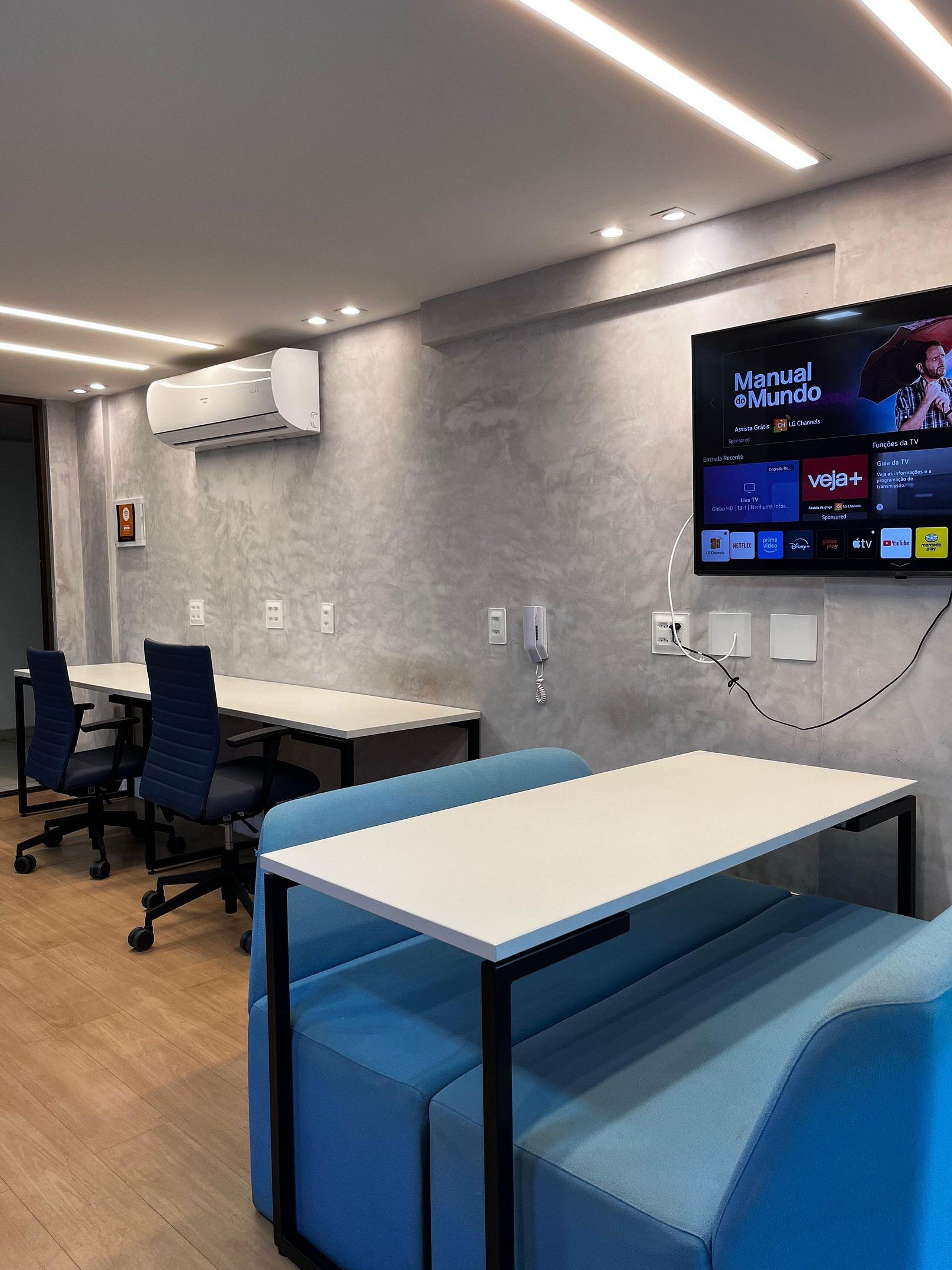 Una sala de conferencias moderna con un televisor de pantalla plana montado en la pared mostrando una guía de televisión. Hay dos mesas con sillas, un sofá azul y un equipo de aire acondicionado en la pared.