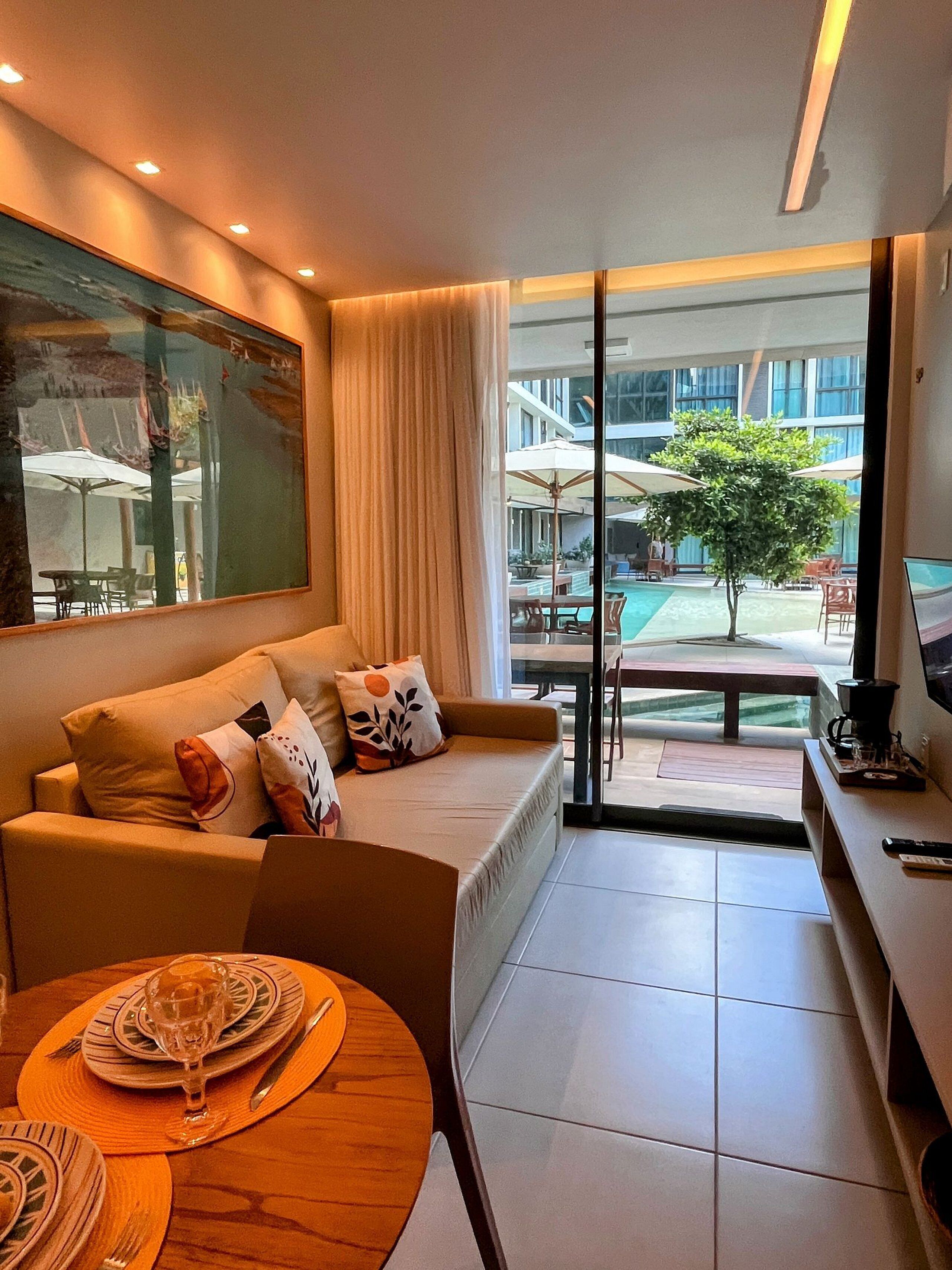 Una sala de estar moderna con un sofá, una mesa de comedor dispuesta con platos y cubiertos, y vistas a un área de piscina al aire libre.