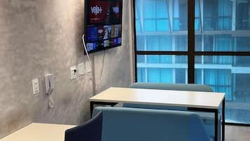 Un espacio de oficina moderno con un escritorio grande, una zona de estar acolchada en azul, un televisor montado que muestra 'veja+' en la pantalla y un ventanal con vistas a un edificio.