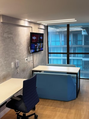 Un espace de bureau moderne doté d’un grand bureau, d’un coin salon bleu avec des coussins moelleux, d’un téléviseur fixé au mur qui affiche « veja+ » à l’écran, et d’une grande fenêtre offrant une vue sur un immeuble.