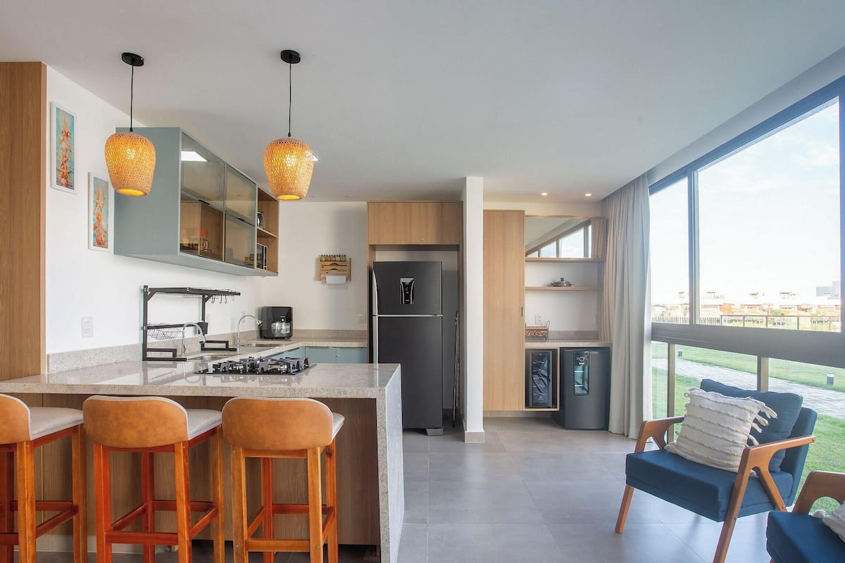 A modern kitchen with a large island, bar stools, a refrigerator, and a view through large windows.