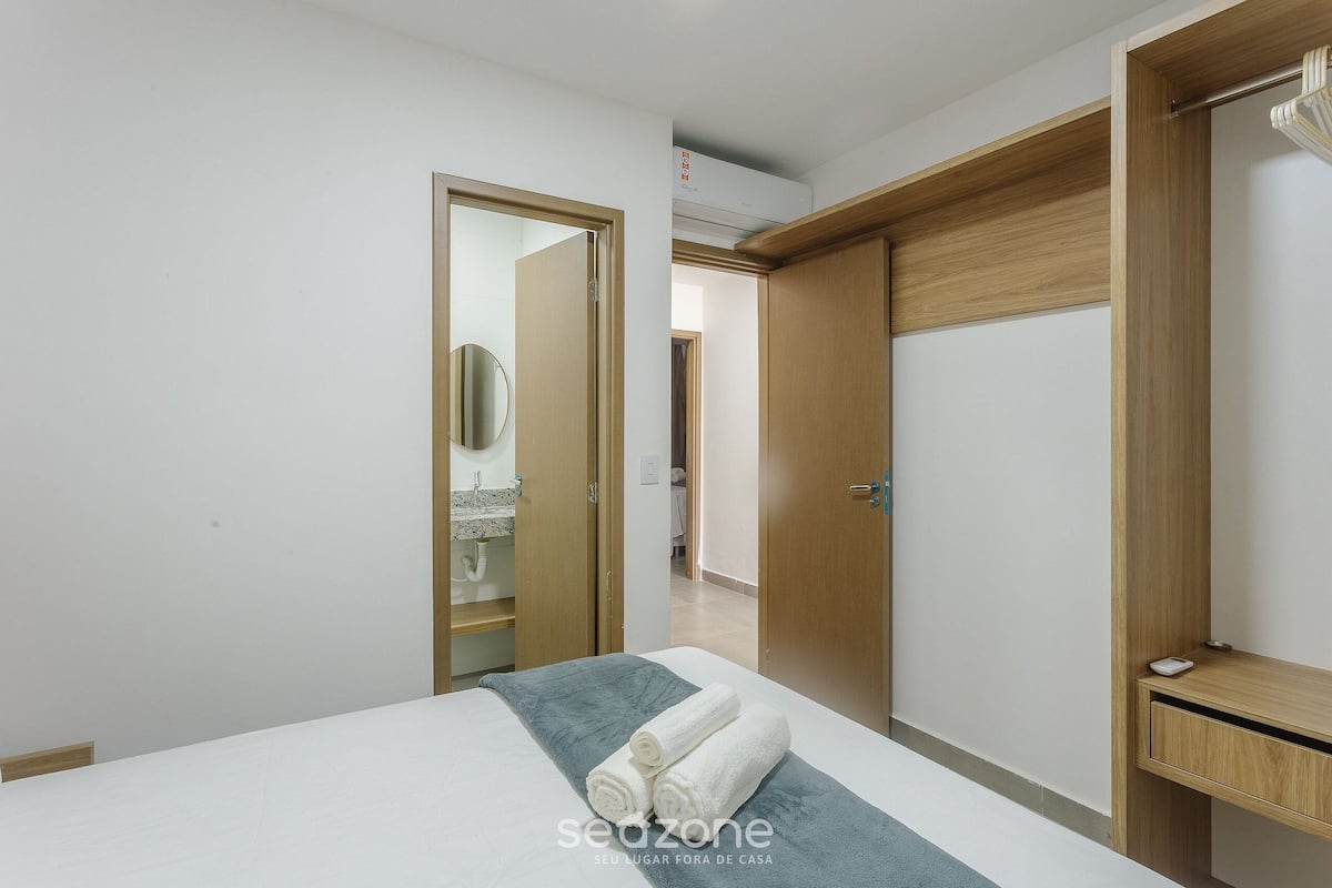 A hotel room with a bed, a wooden wardrobe, a bathroom with a mirror, and a ceiling air conditioner.