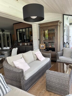 Uma sala de estar moderna com um sofá de vime cinza, um candeeiro pendente escuro e uma estante de madeira repleta de livros.
