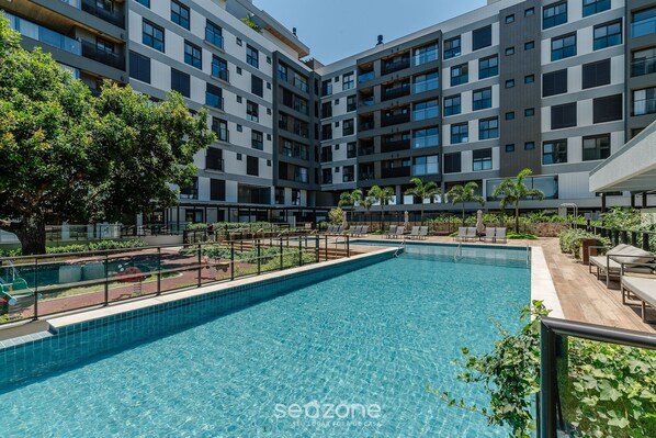 A modern building with a swimming pool, surrounded by greenery and outdoor seating.