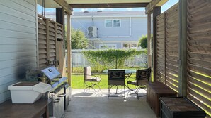 A covered patio area with a grill, outdoor furniture, and a view of a house in the background.