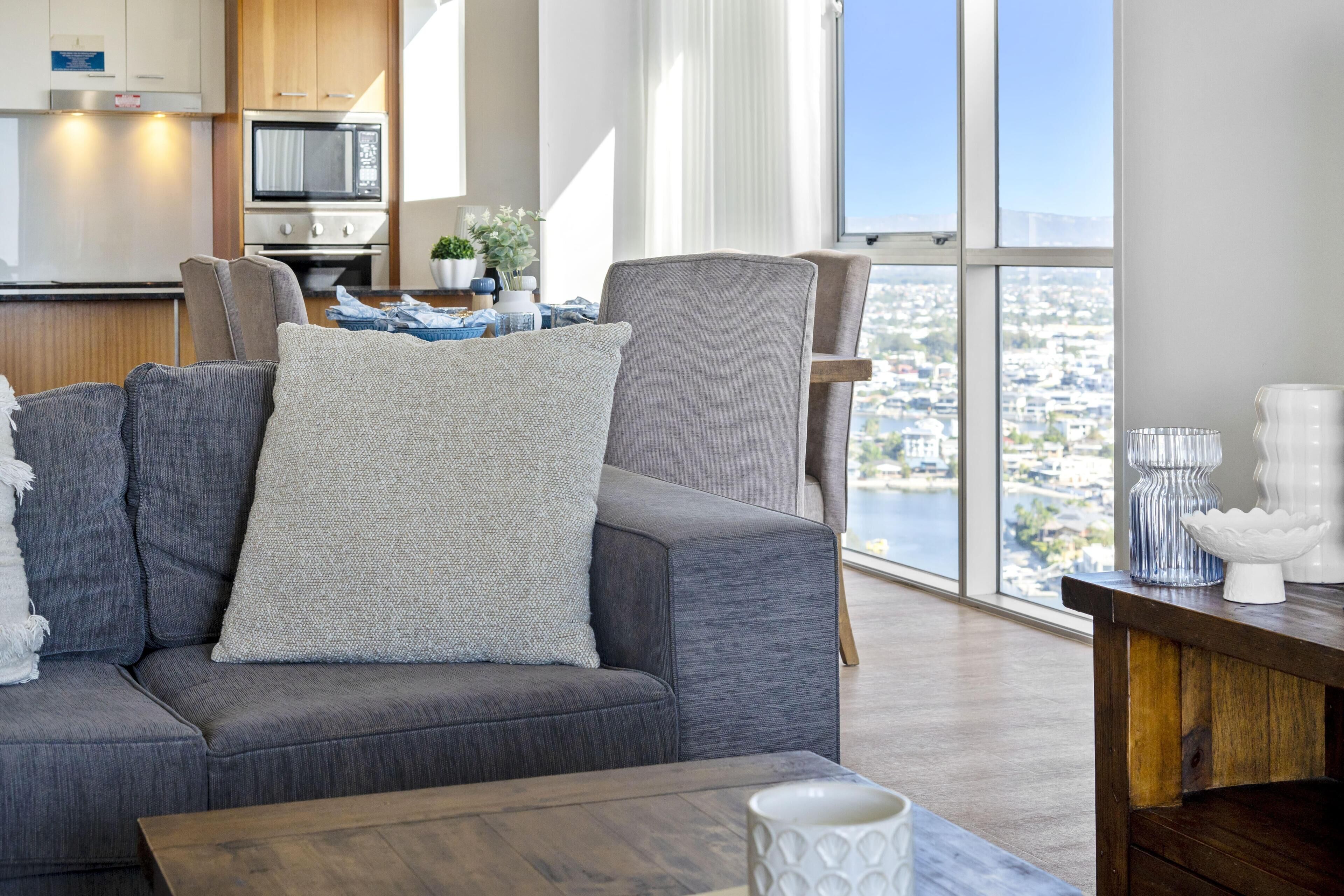 A modern living room with a gray sofa, a wooden coffee table, and a view of a cityscape through large windows.