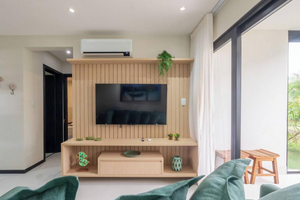 A modern living room with a flat-screen TV mounted on a wooden wall, a built-in air conditioner, and a small shelf with decorative items.
