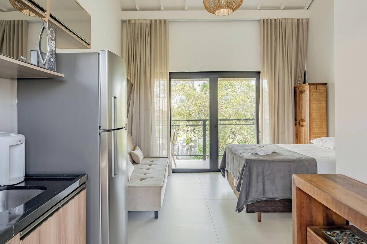 A compact hotel room with a kitchenette, a bed, a bench, and a view through a glass door.