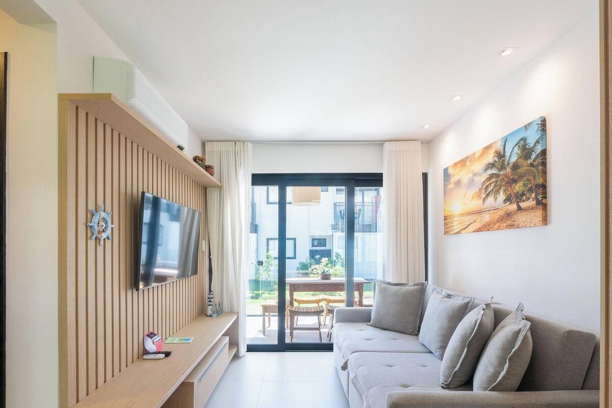 A modern living room with a flat-screen TV, a wooden panel wall, a sofa, a painting, and a view of the outdoors.