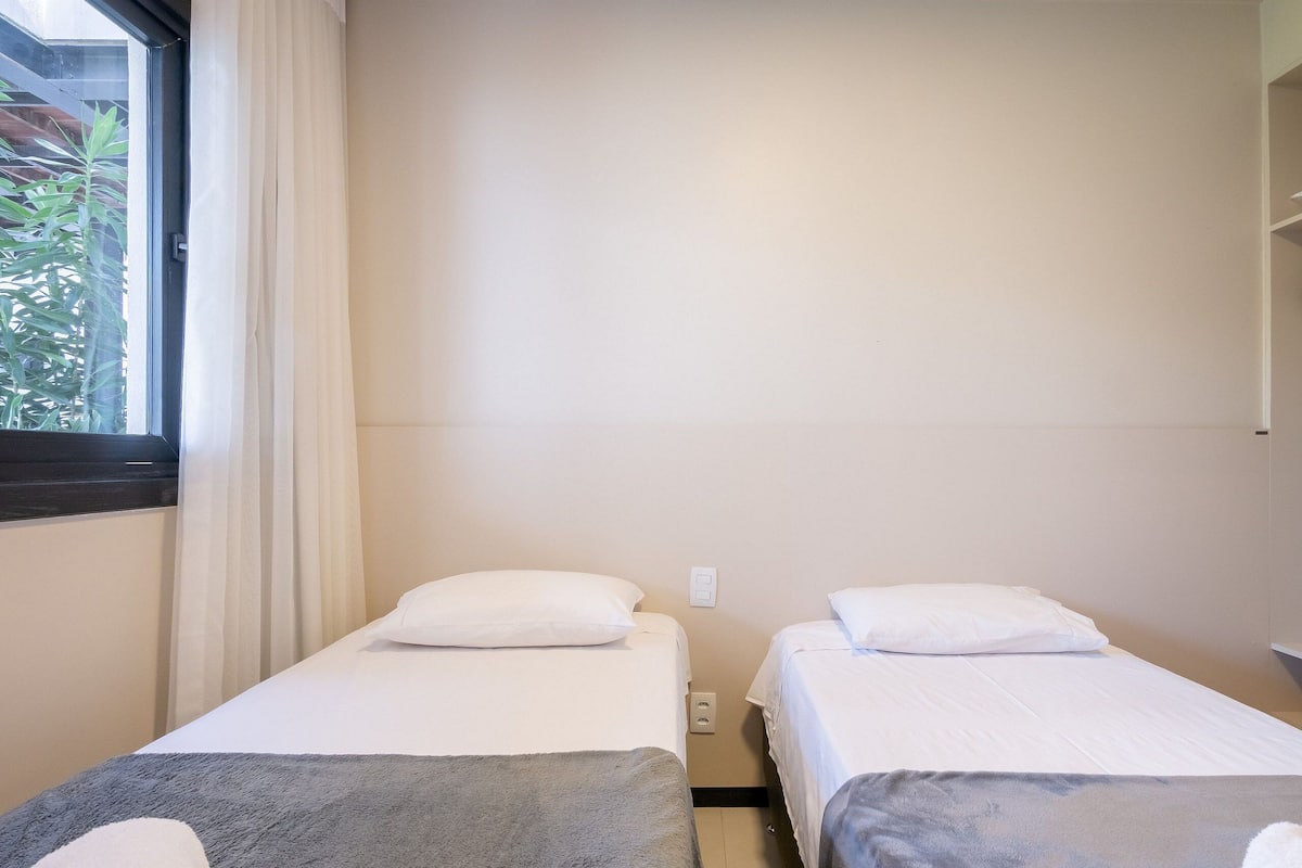 A room with two beds, a window with curtains, and a view of greenery outside.