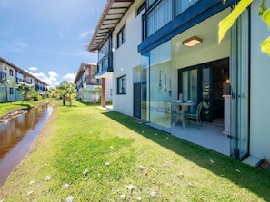 A modern building with a glass door entrance, a small outdoor seating area, and a narrow water channel. - VLP - Apartments Close to Praia do Forte/ba (Mata de São João)
