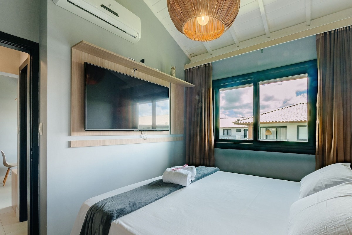 A hotel room with a bed, a television mounted on the wall, a window with a view of buildings, and a ceiling fan.