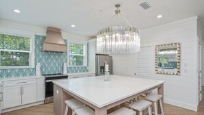 Une cuisine moderne avec des meubles blancs, un îlot central doté de tabourets, un grand lustre et un pare-feu décoratif.