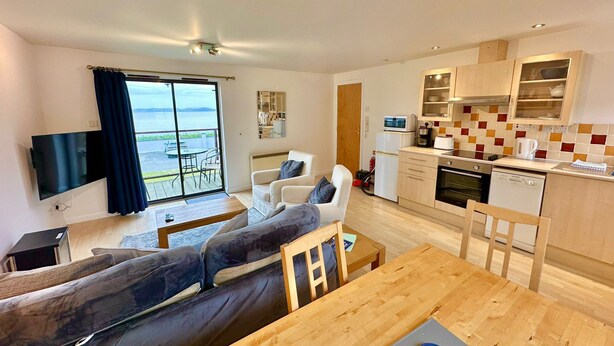 Een modern appartement met een woongedeelte, eethoek en keuken. Het heeft een groot raam met uitzicht op zee, een flatscreen-tv, een bank, een eettafel en een keuken met moderne apparatuur.