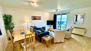 Een moderne woonkamer met een houten eettafel, twee banken, een televisie en een groot raam met uitzicht op zee.