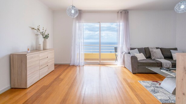 Un moderno salón con suelo de madera, un sofá gris, un armario blanco y vistas al mar a través de amplios ventanales.
