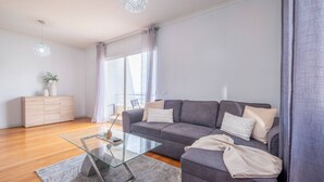 Una sala de estar moderna con un sofá gris, una mesa de centro de vidrio, un mueble de madera y un amplio ventanal con cortinas.