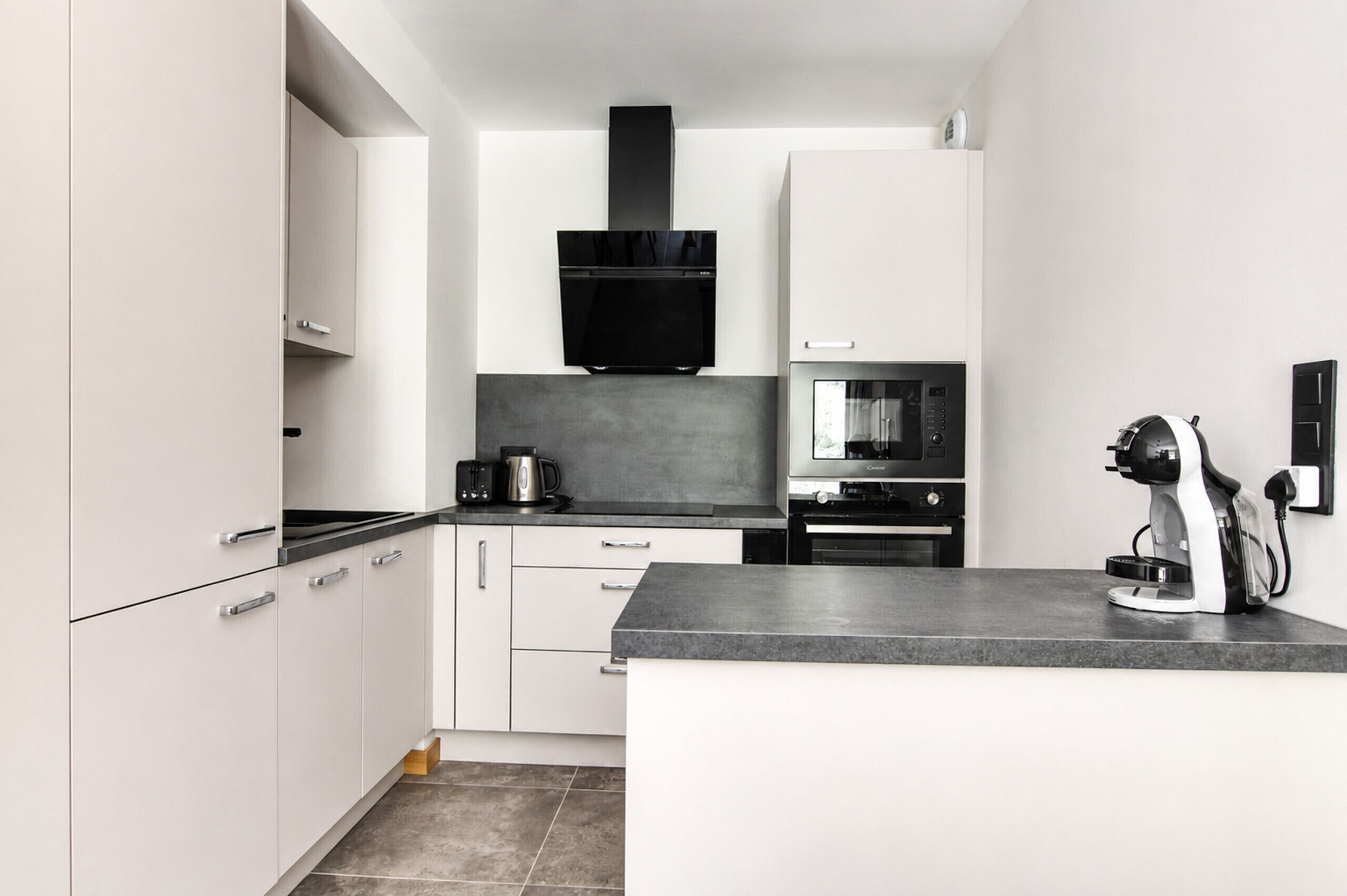 A modern kitchen with white cabinets, a black range hood, a coffee machine, and a microwave.