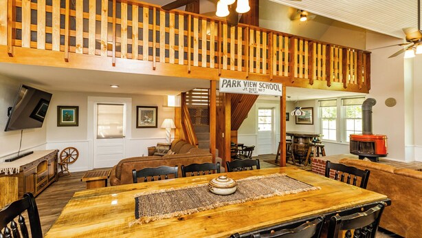 A spacious dining area with a long wooden table, multiple chairs, and a high ceiling with exposed beams. A staircase leads to an upper level with a wooden railing.