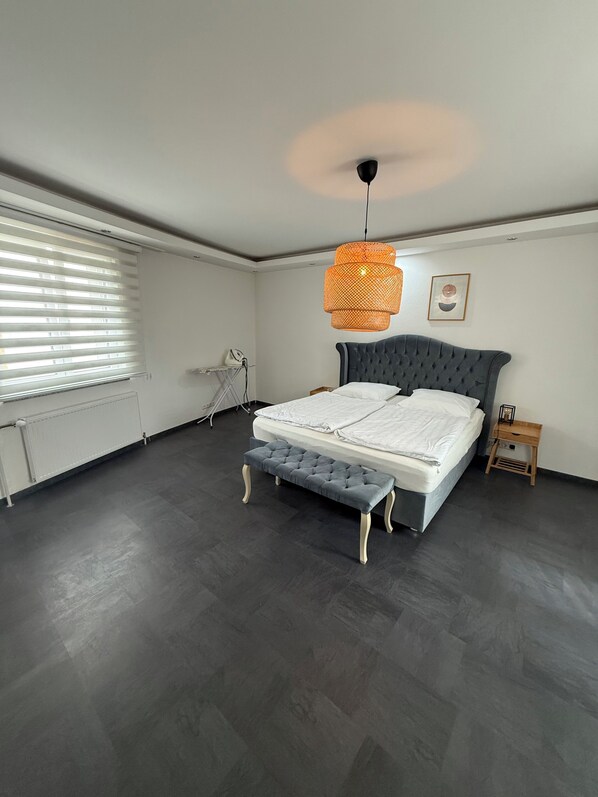 A bedroom with a bed, a bench, a heater, a radiator, and a window with blinds.