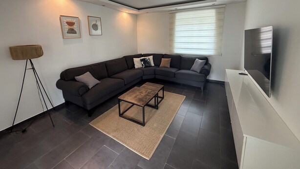 A modern living room with a dark grey sectional sofa, a wooden coffee table, a floor lamp, and a flat-screen TV mounted on the wall.