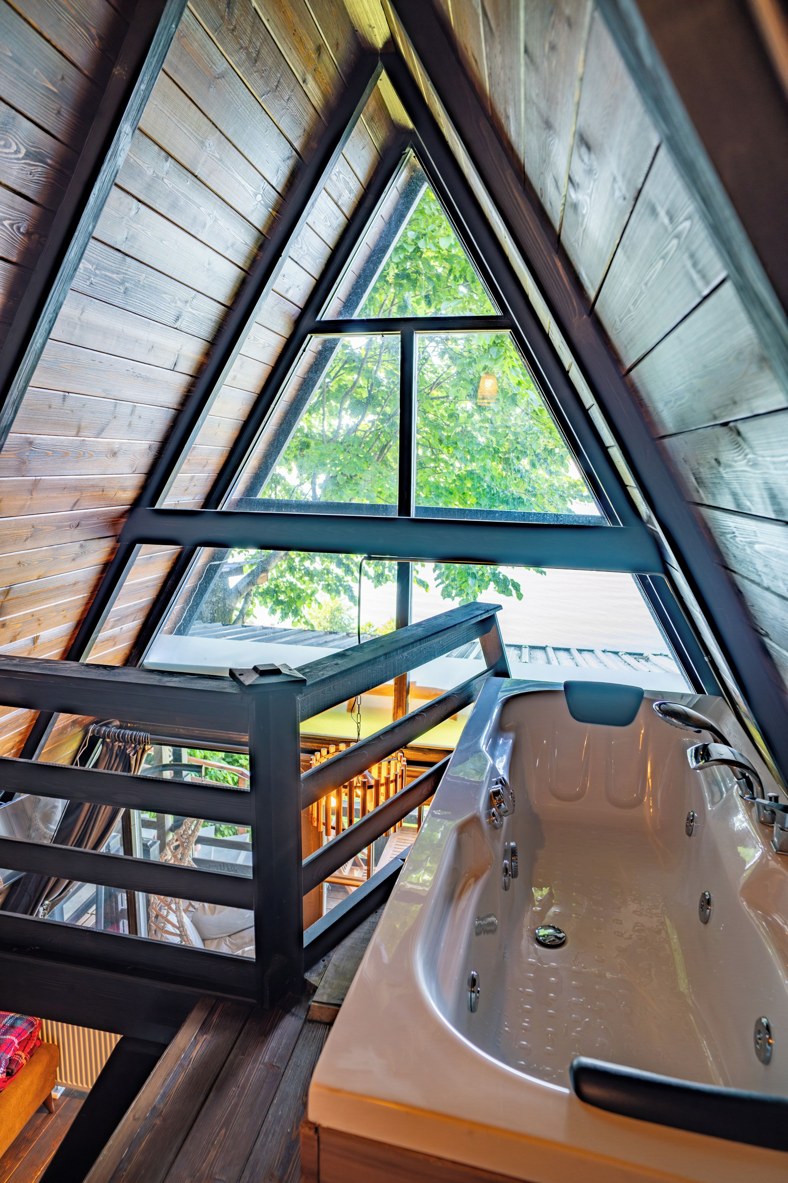 A wooden interior with a hot tub, a wooden staircase, and a view through a triangular window.
