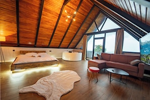 A spacious room with a high ceiling, a bed with a headboard, a seating area with a couch and a small table, and a large window providing a view outside.