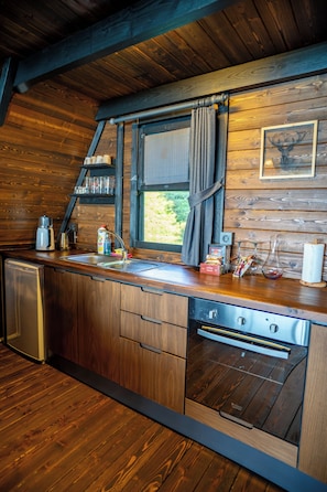 A compact kitchen with wooden cabinetry, a built-in dishwasher, and a window with curtains.
